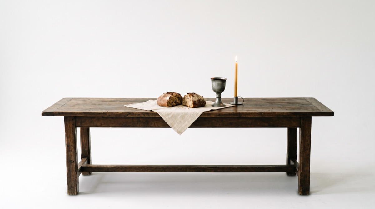 A broken loaf, a pewter chalice, and one lit candle on a long dark table in a pure white room.